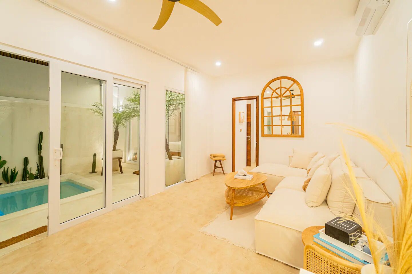 A modern living room with a glass door leading to a pool area, a round wooden table, and a cozy sofa with cushions.