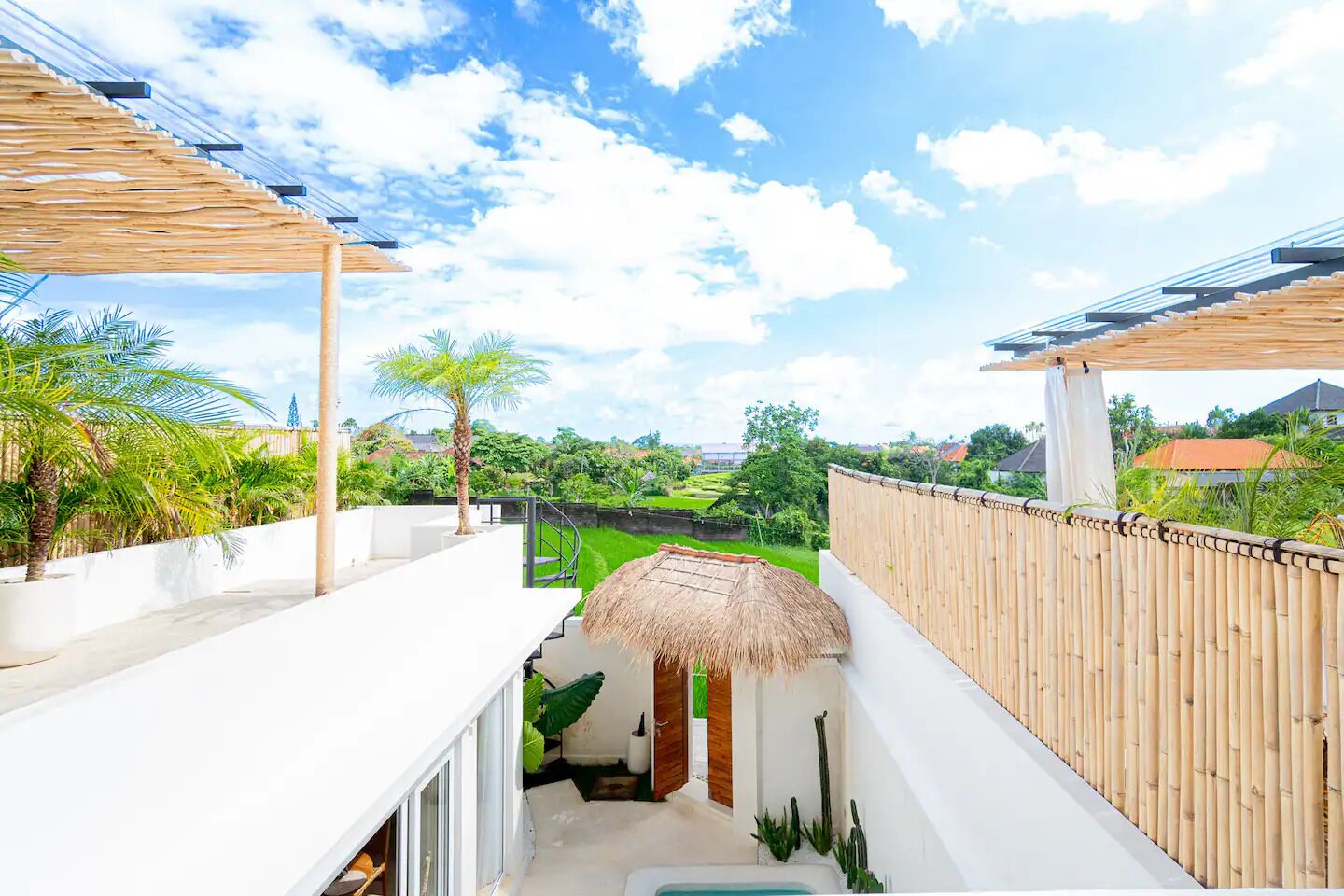 A modern outdoor balcony with a thatched roof, white walls, and a view of a lush garden and distant buildings.