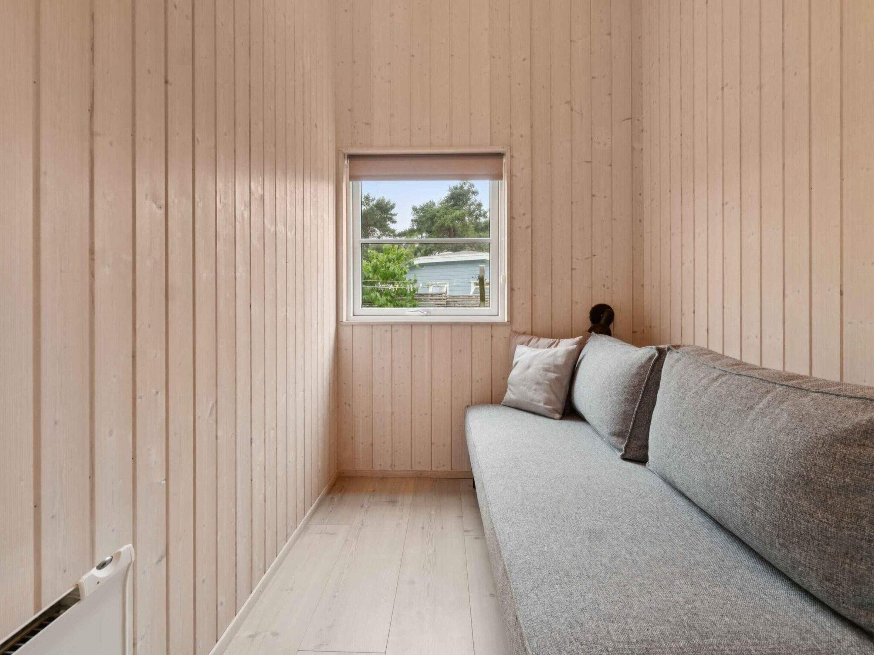 A room with wooden walls, a window with a view of greenery and a building, a grey sofa with pillows, and a radiator on the floor.
