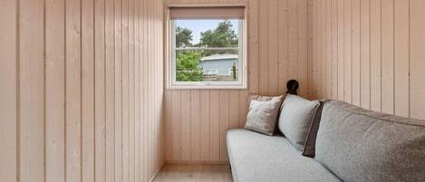 A room with wooden walls, a window with a view of greenery and a building, a grey sofa with pillows, and a radiator on the floor.