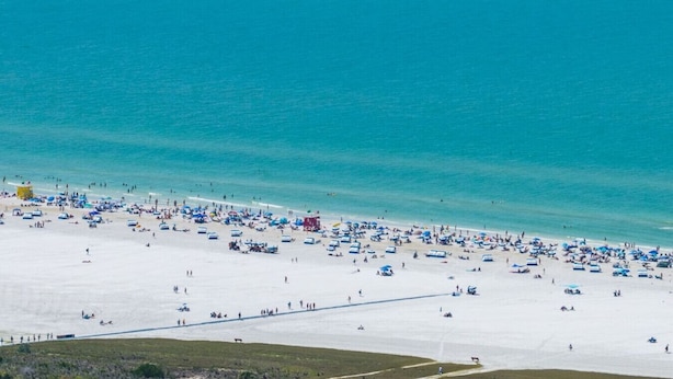 A beach with numerous umbrellas and people, a clear blue sea, and a boat creating a wake. - The Pelican on Siesta (Siesta Key)