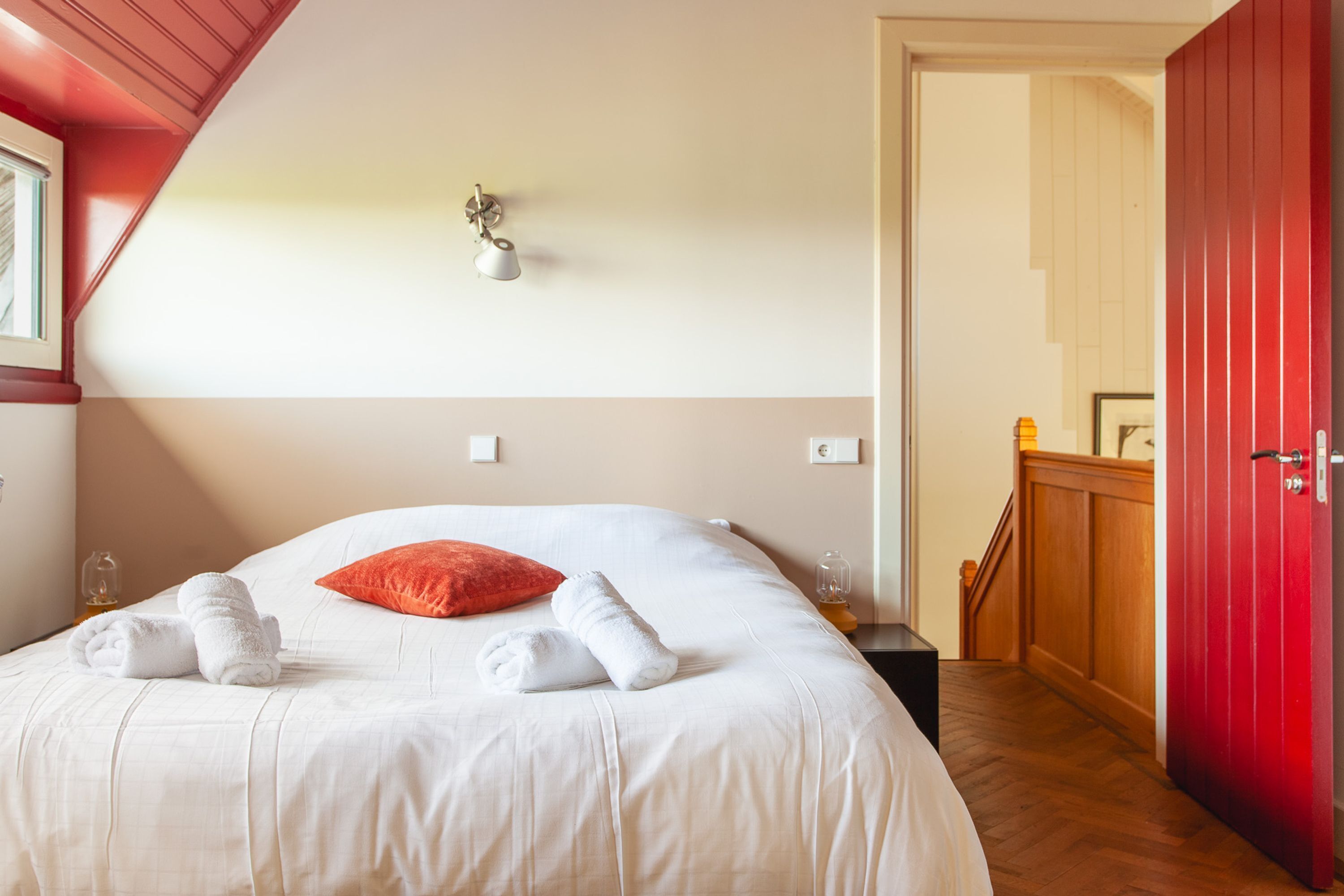A neatly made bed with white linens and a red pillow, a bedside table with a lamp, and a wooden staircase leading to a red door.