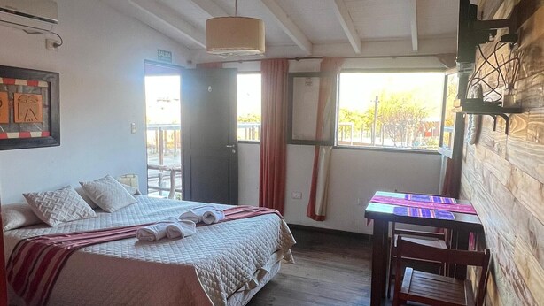 Una habitación con dos camas, piso de madera, una mesa con sillas, una ventana con cortinas y una puerta.