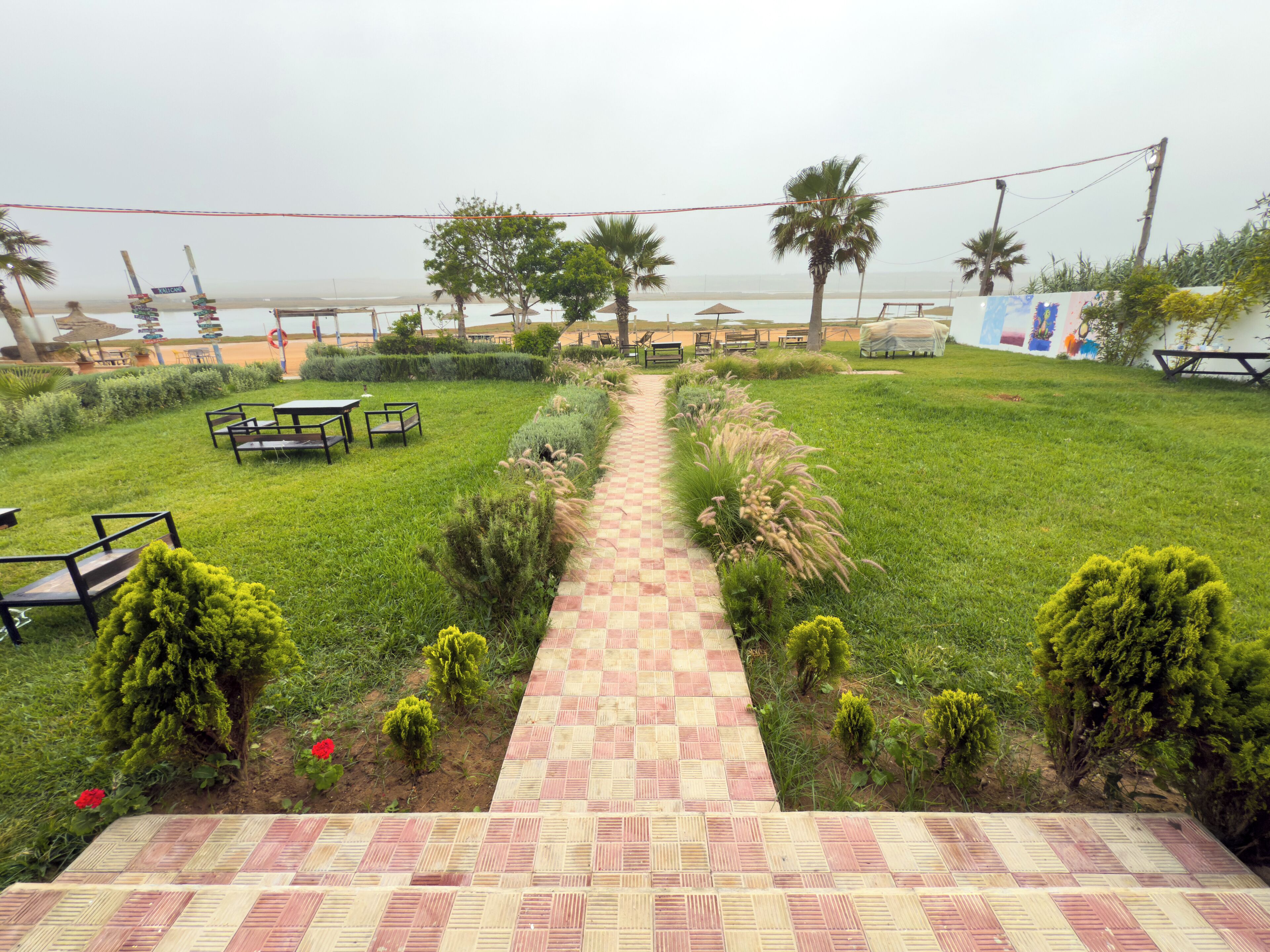 A garden pathway leading to outdoor seating with tables and chairs, surrounded by greenery and palm trees.