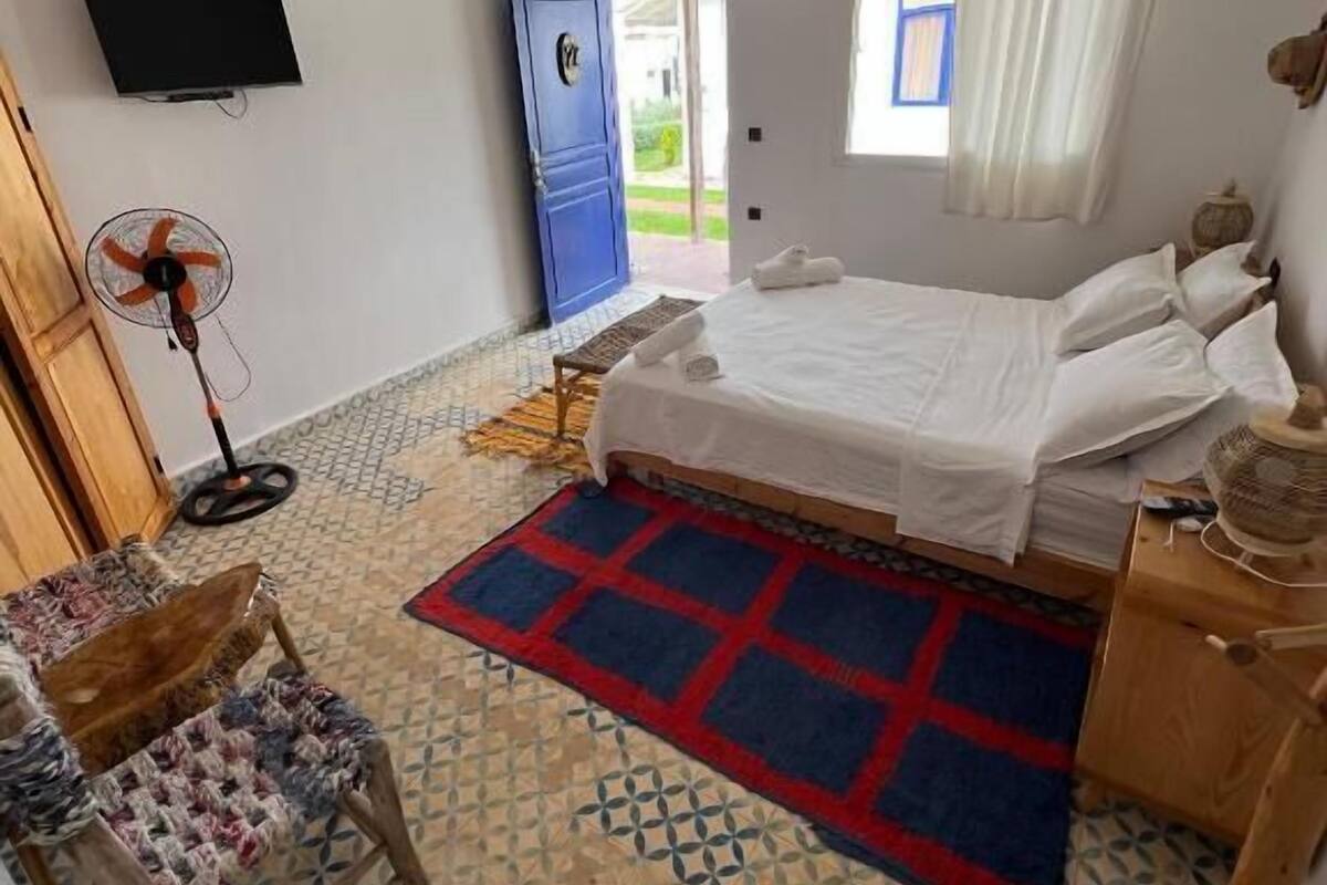 Ein Schlafzimmer mit Bett, Fernseher, Ventilator, einem Fenster mit Vorhängen und Blick ins Freie.