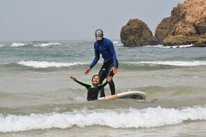 Eine Person im Neoprenanzug hilft einer anderen beim Surfen im Meer, im Hintergrund sind Felsen zu sehen.