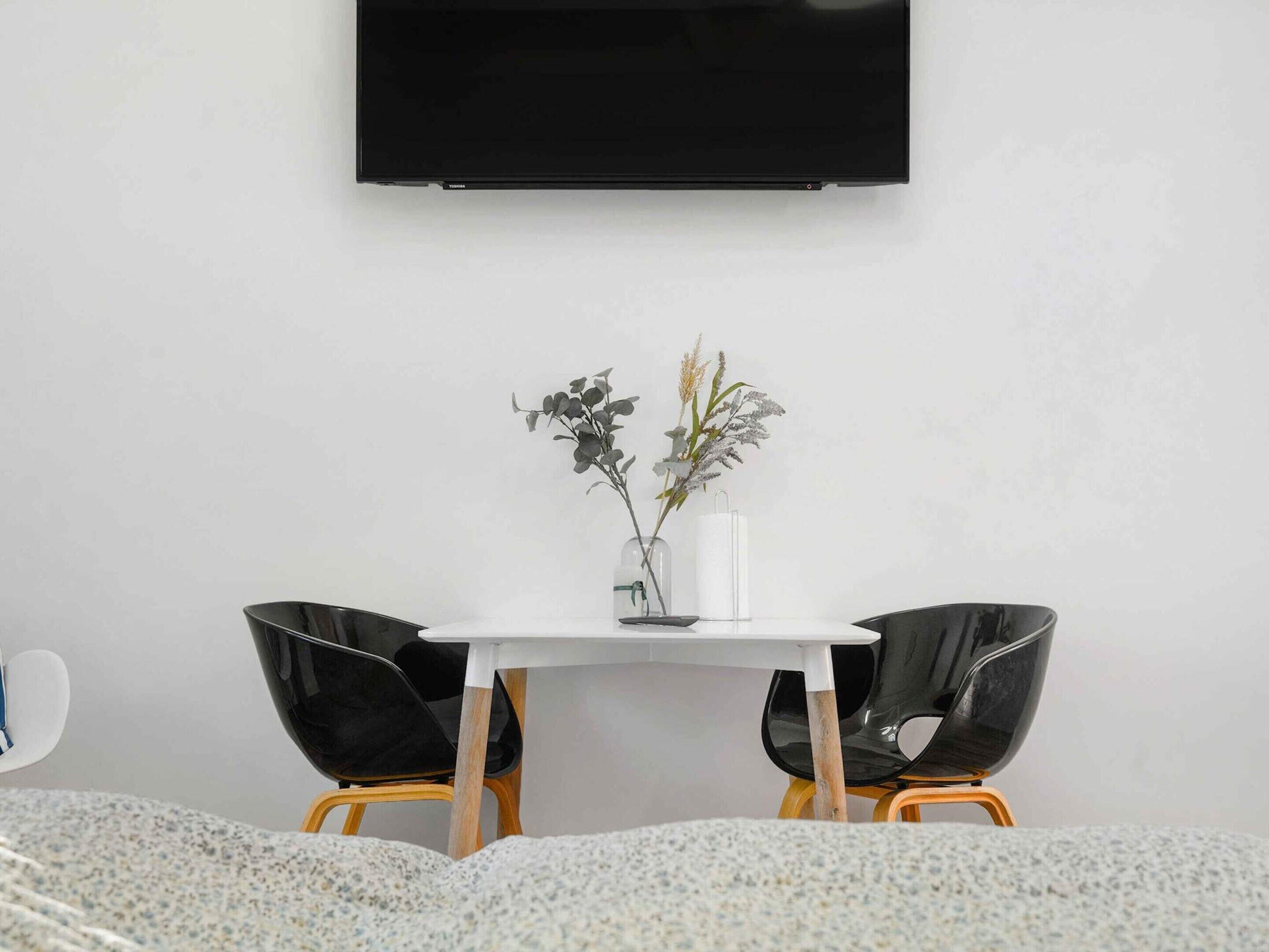 Ein modernes Zimmer mit einem weißen Tisch, zwei schwarzen Stühlen und einem an der Wand befestigten Flachbildfernseher.