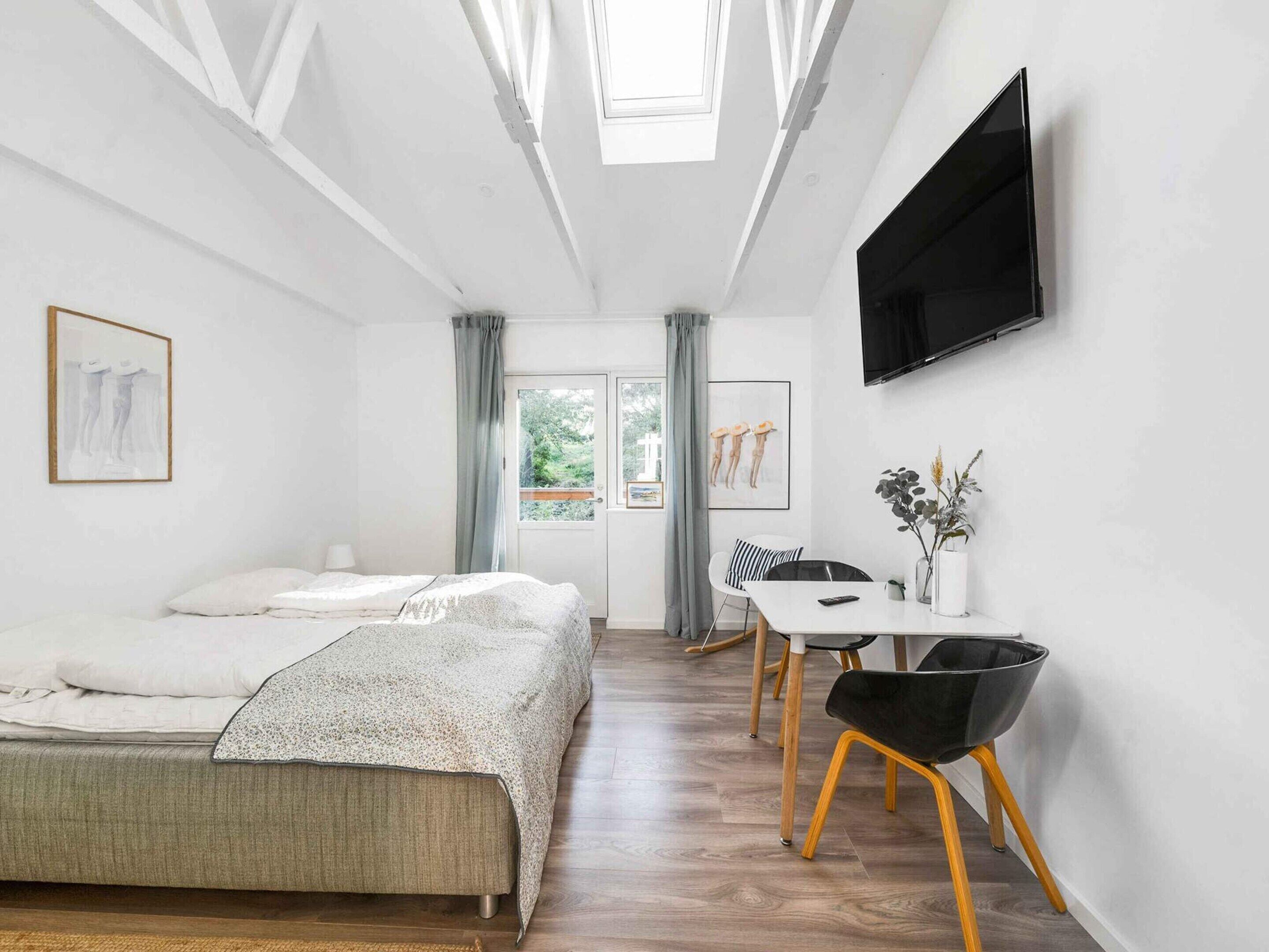 A modern room with a bed, a dining table with chairs, a TV mounted on the wall, and a window with curtains.