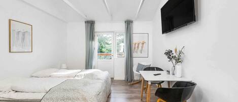 A modern room with a bed, a dining table with chairs, a TV mounted on the wall, and a window with curtains.
