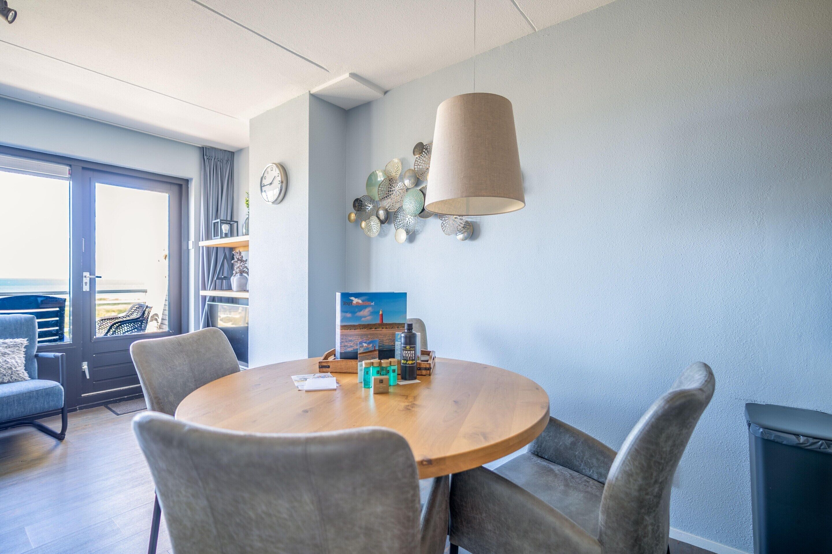A dining area with a round wooden table, four grey upholstered chairs, a hanging lamp, and a decorative wall piece.