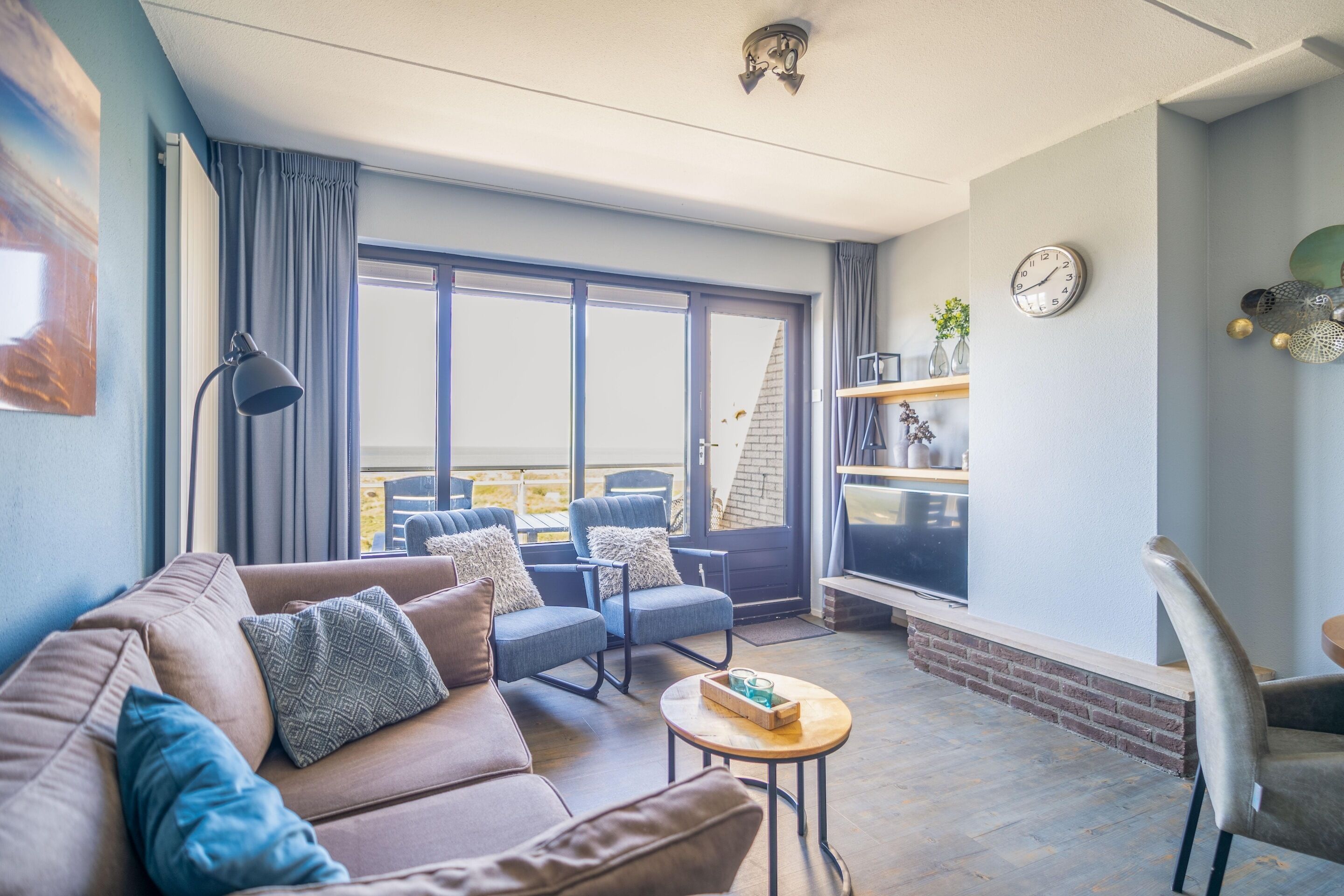 A modern living room with a brown sofa, two blue armchairs, a small round table, and a large window with curtains.