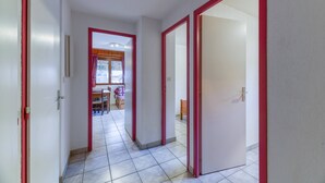 Un couloir avec des portes à cadre rouge, un sol carrelé et une vue sur un coin repas avec une table et des chaises.