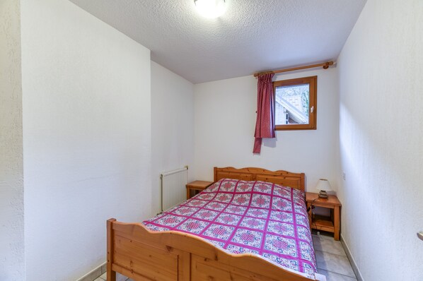 Un dormitorio pequeño con cama de madera, una mesita de noche, un ventanal con cortina roja y un radiador.