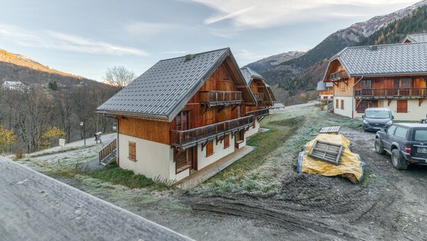 Un chalet en bois doté d’un balcon, un sentier de terre et une vue sur les montagnes en arrière-plan.