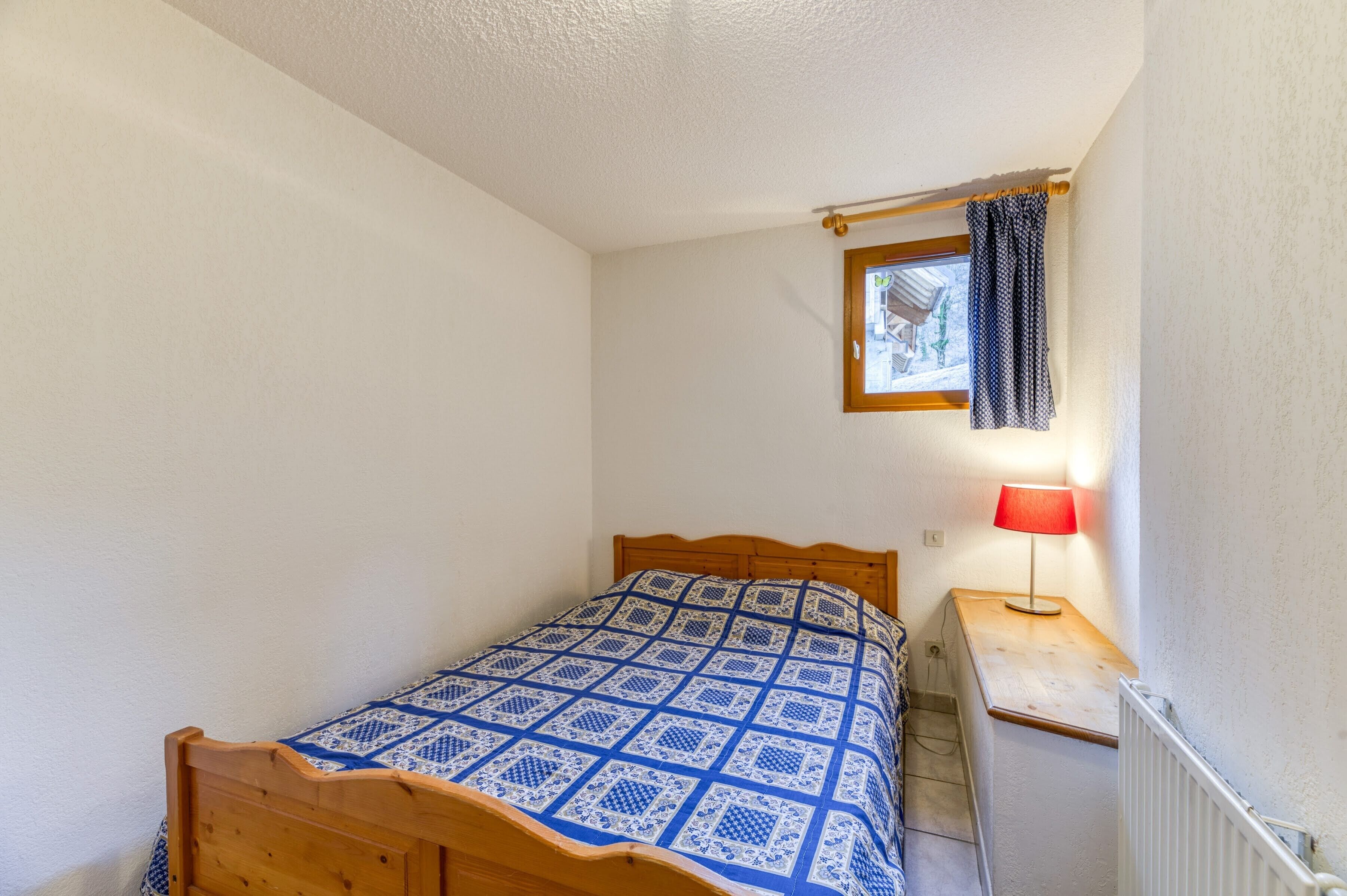 A small bedroom with a wooden bed, a nightstand with a lamp, a window with curtains, and a radiator.