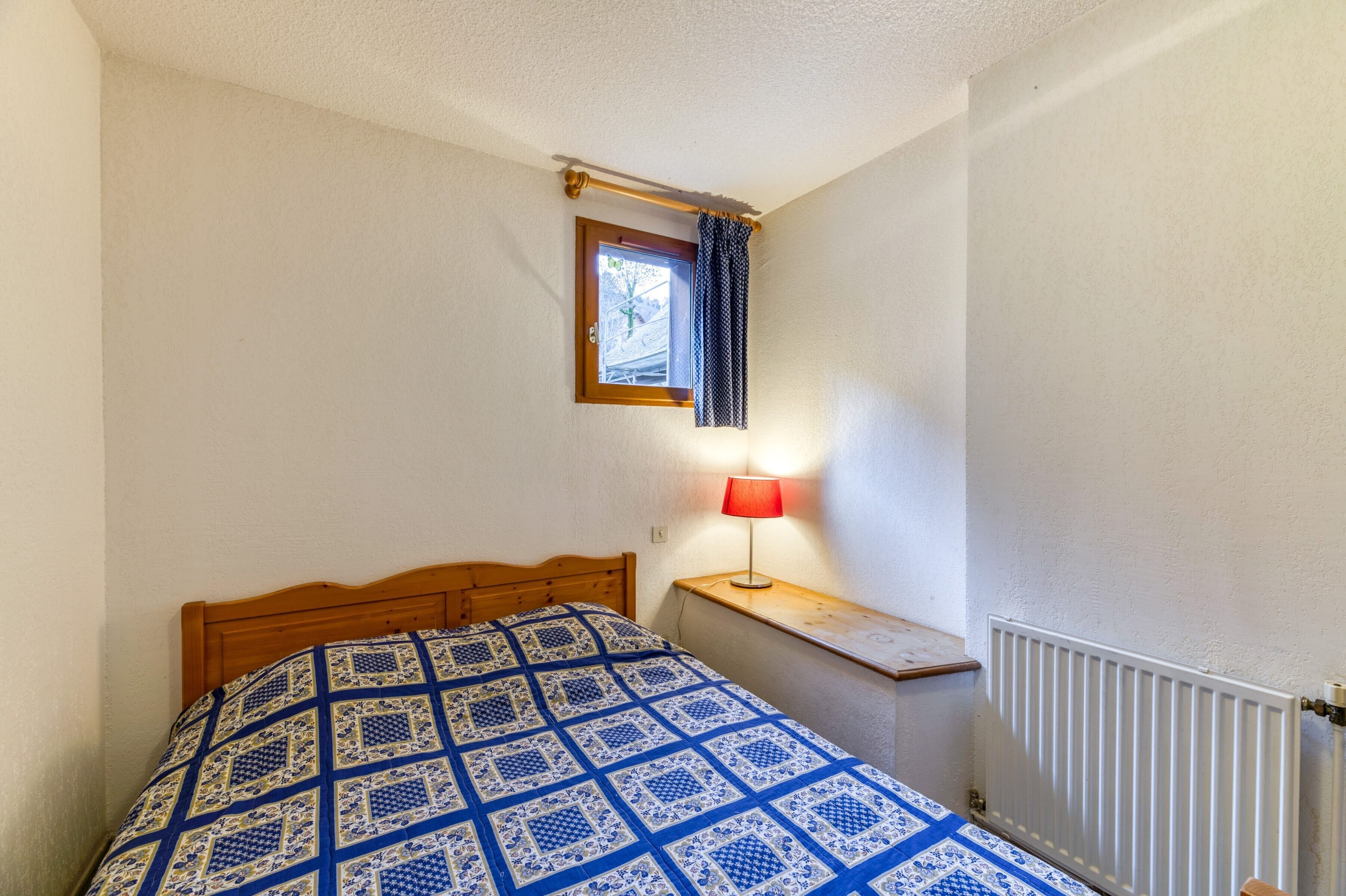 Quarto pequeno com cama de madeira, abajur vermelho sobre uma mesa de cabeceira, janela com cortina e radiador.