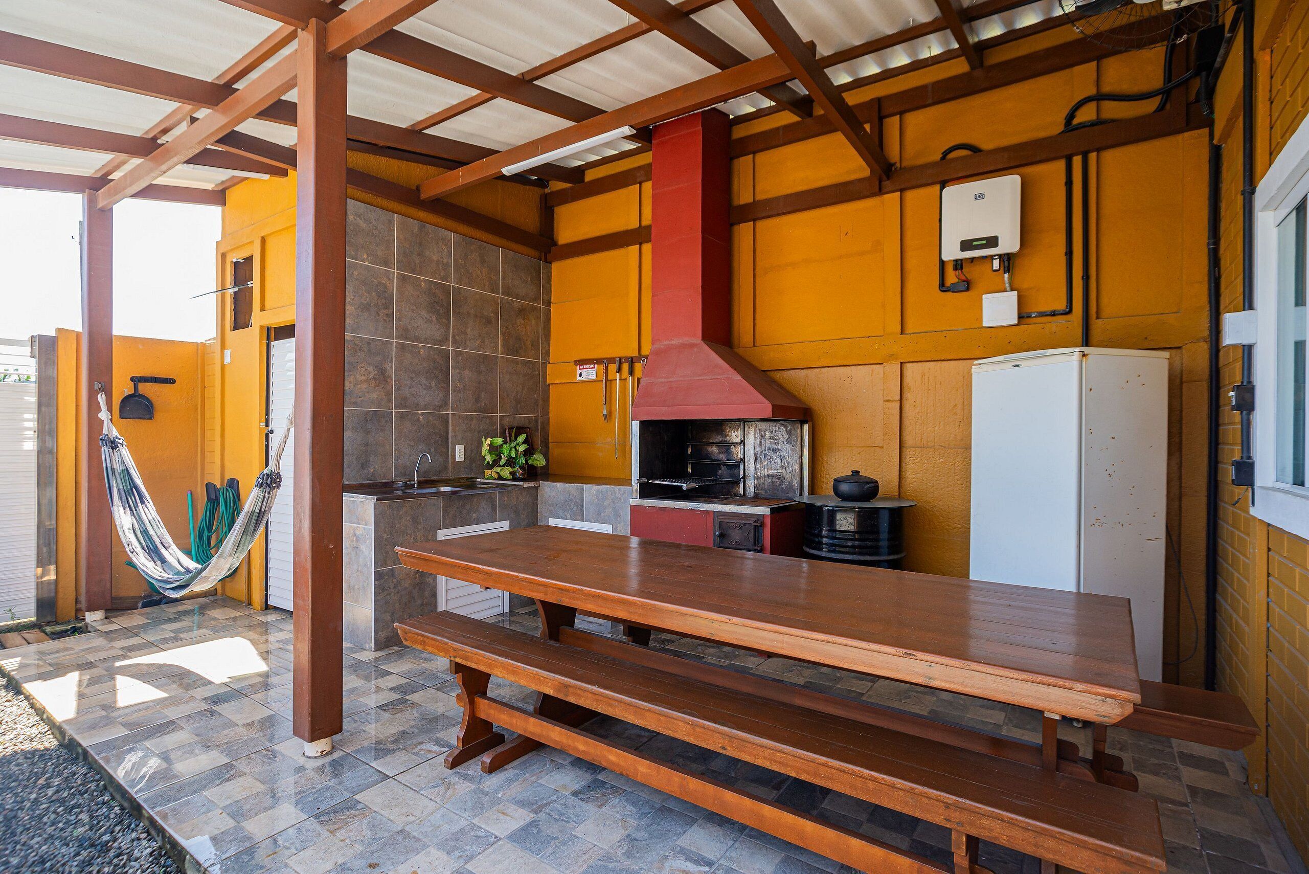 A covered outdoor dining area with a wooden table and benches, a hammock, and a kitchen area with a stove and refrigerator.