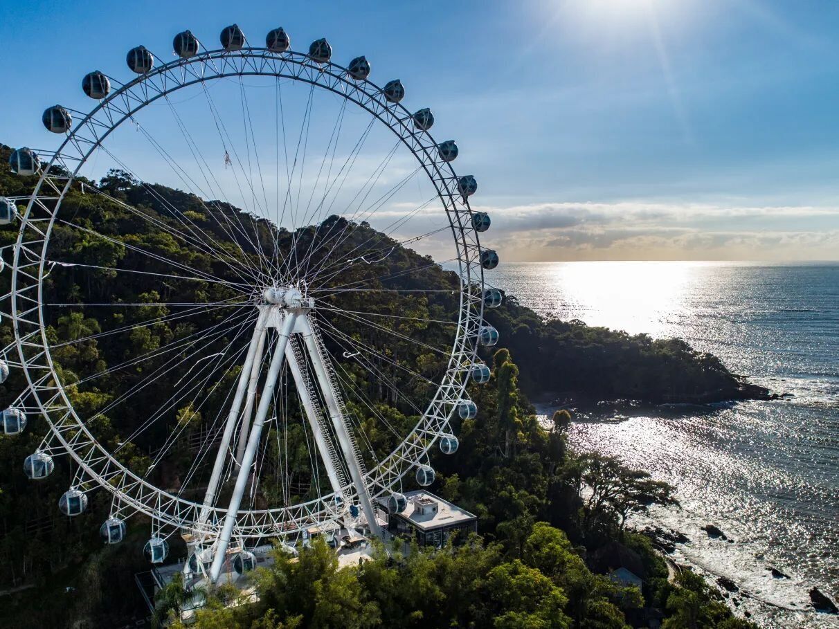 A large Ferris wheel with gondolas, surrounded by lush greenery and overlooking a coastal area.