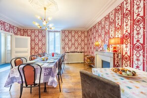 Une salle à manger avec une longue table dressée pour six personnes, un lustre, un papier peint à motifs et une cheminée agrémentée d’une lampe de table.