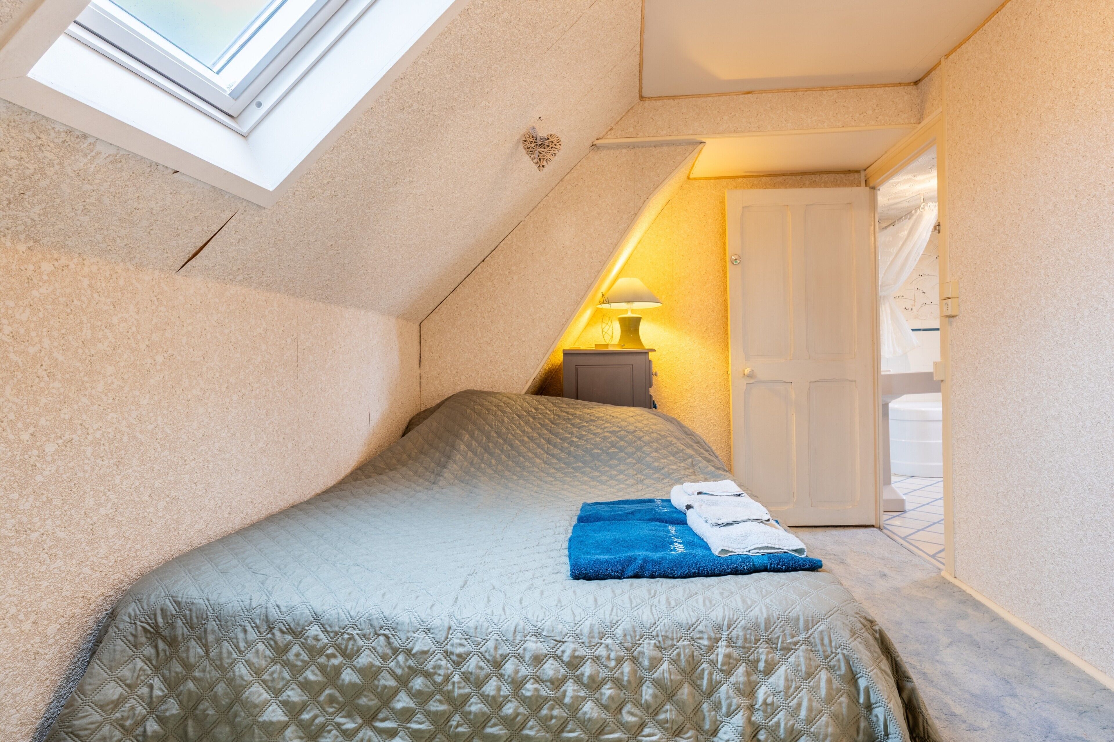 A bedroom with a sloped ceiling, a bed with a quilted cover, a nightstand with a lamp, and a door leading to another room.