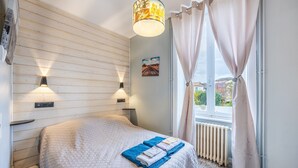 Une chambre à coucher avec un lit, deux lampes fixées au mur, une fenêtre avec des rideaux, un radiateur et un tableau représentant un paysage urbain accroché au mur.
