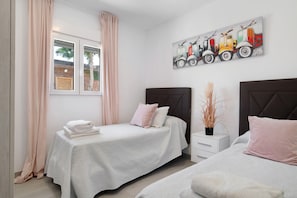 Une chambre avec deux lits, une fenêtre avec des rideaux, un tableau représentant des voitures anciennes accroché au mur et une plante sur la table de chevet.