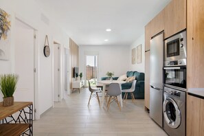 Een moderne keuken met een inbouwoven en magnetron, een eethoek met een ronde tafel en stoelen, en een zithoek met een bank en een plant.