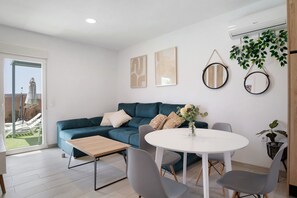 Un salon moderne avec un canapé de couleur turquoise, une table ronde blanche et des chaises grises. Deux œuvres d’art abstraites encadrées ornent le mur, ainsi qu’un miroir suspendu et une plante en pot.