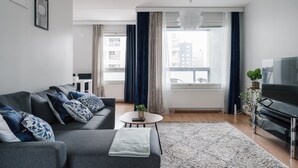 A modern living room with a grey sofa, a coffee table, a flat-screen TV, and a large window with curtains. - Spacious City Retreat with Private Sauna (Helsinki)
