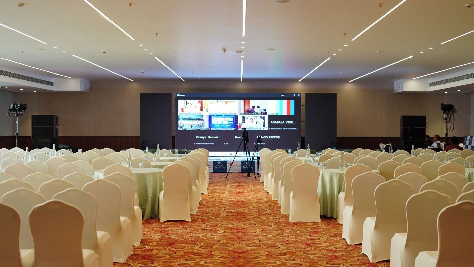 A conference room with rows of white chairs, a large screen at the front, and a carpeted floor.