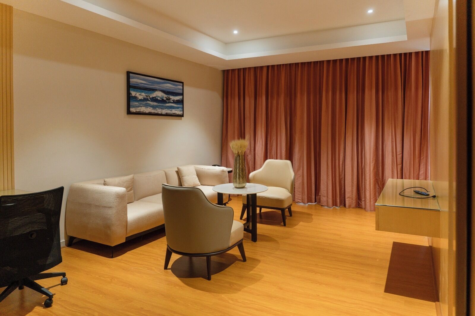 A modern hotel room with a sofa, two armchairs, a small table, a desk with a lamp, and a painting on the wall.