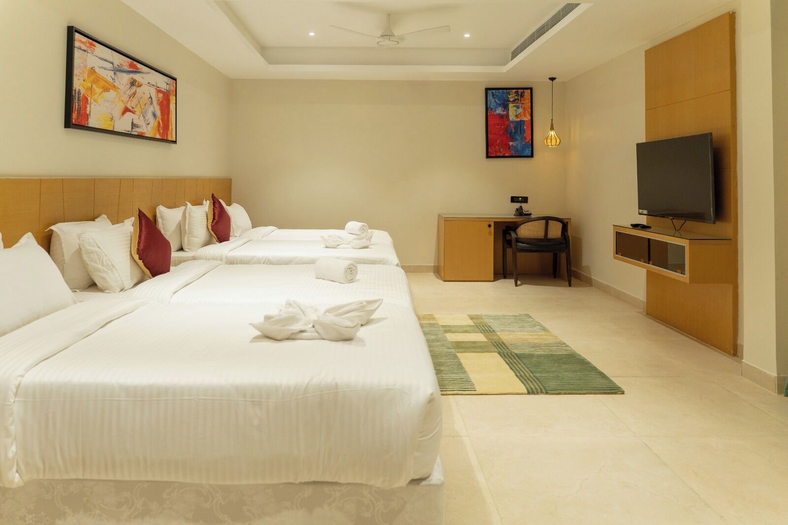 A hotel room with three beds, a desk, a television, and artwork on the walls.