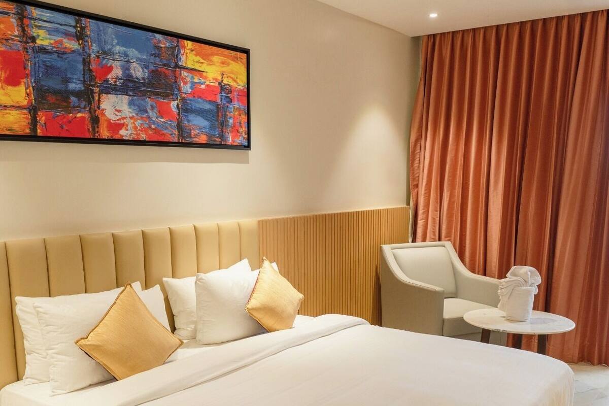 A modern hotel room with a large bed, a bedside table, a chair, and a small table with a vase.