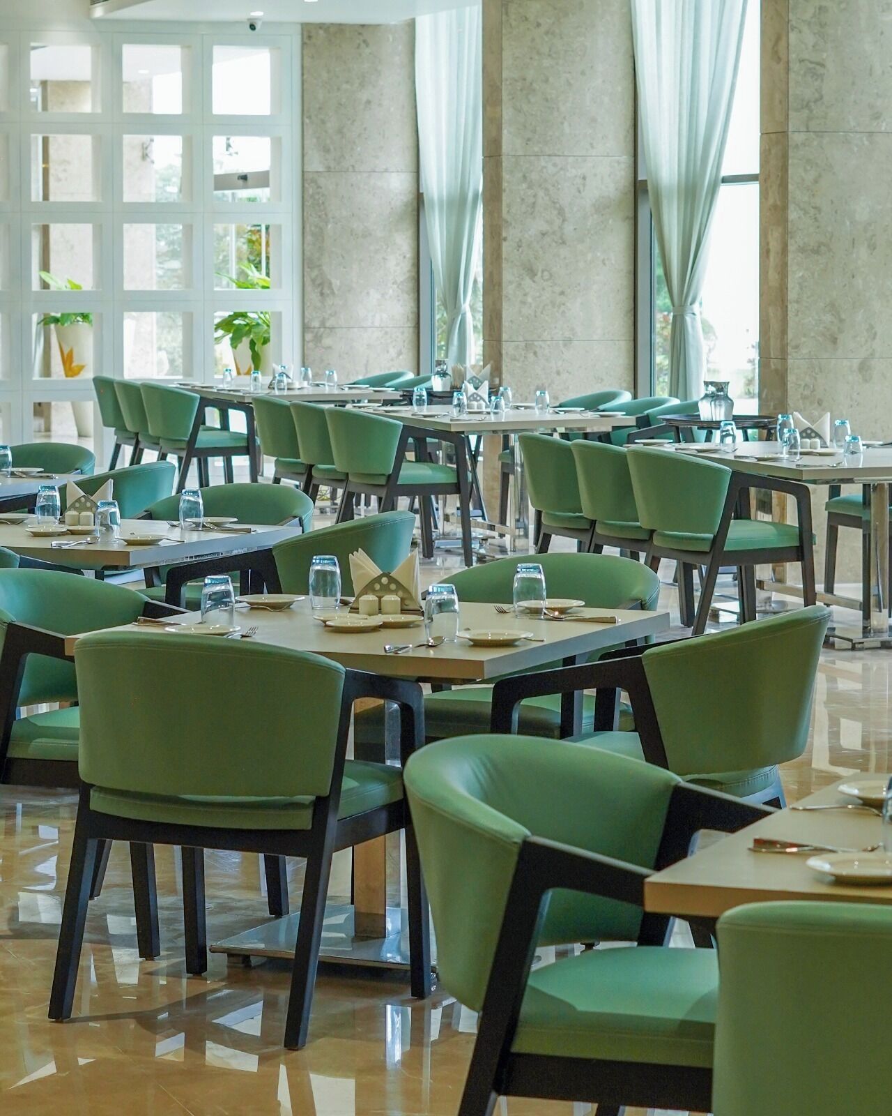 Un comedor amplio con varias mesas y sillas, ventanas grandes con cortinas y plantas en macetas.