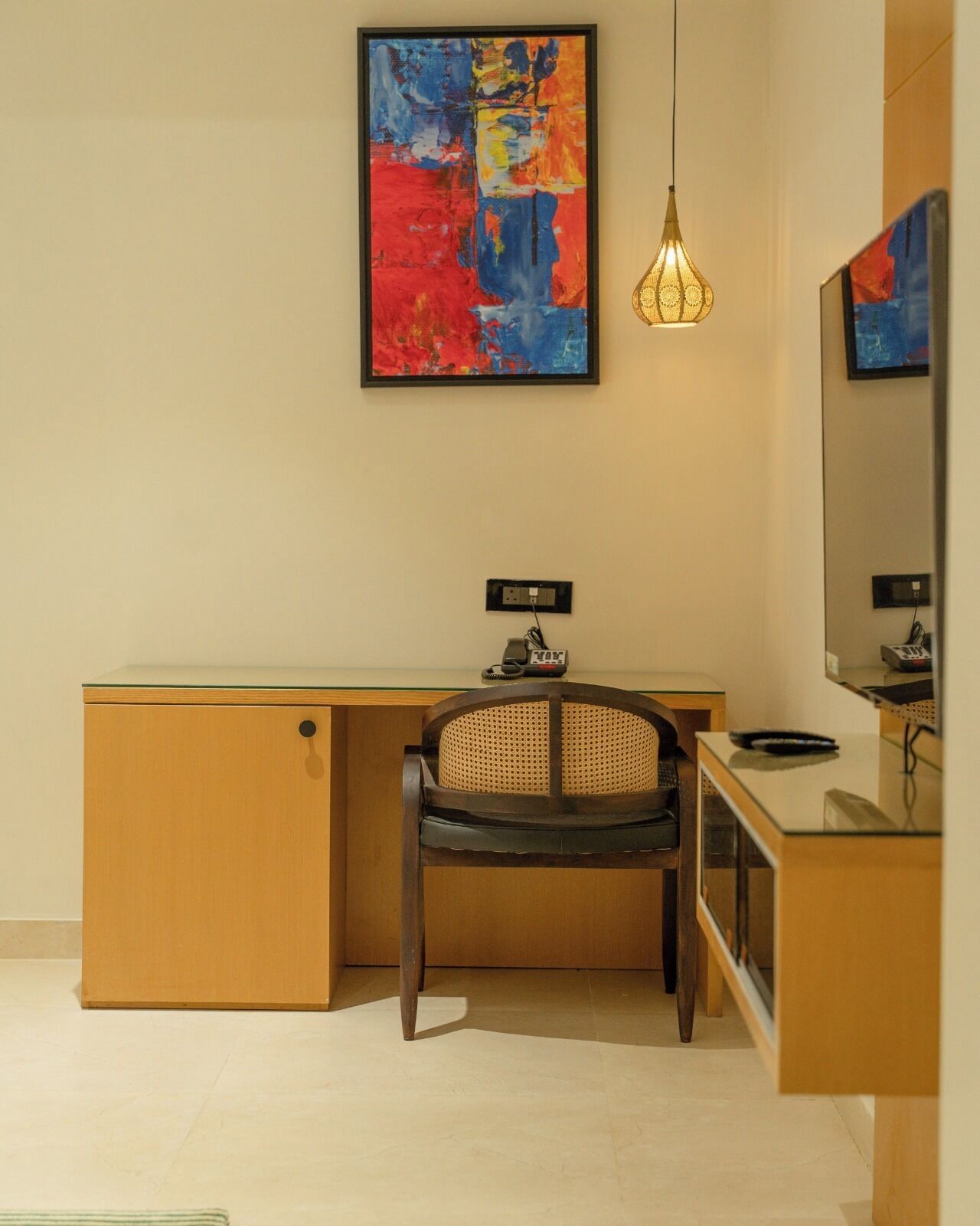 A modern bathroom with a wooden vanity, a chair with a woven backrest, a large mirror, and a colorful abstract painting on the wall.