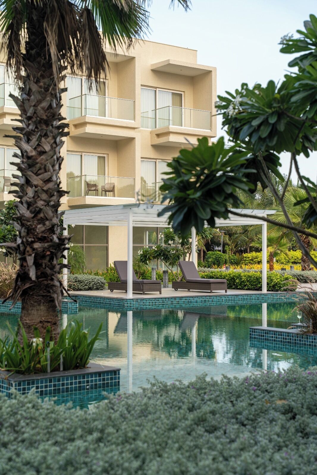 A modern hotel with a swimming pool, lounge chairs, and lush greenery.