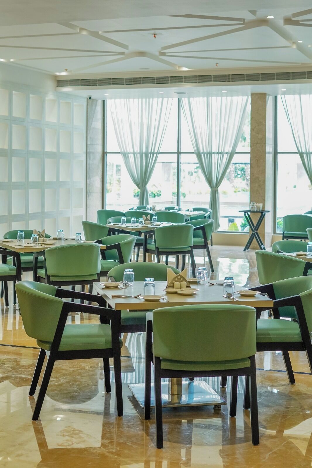 A dining area with green chairs and tables, large windows with curtains, and a modern ceiling design.