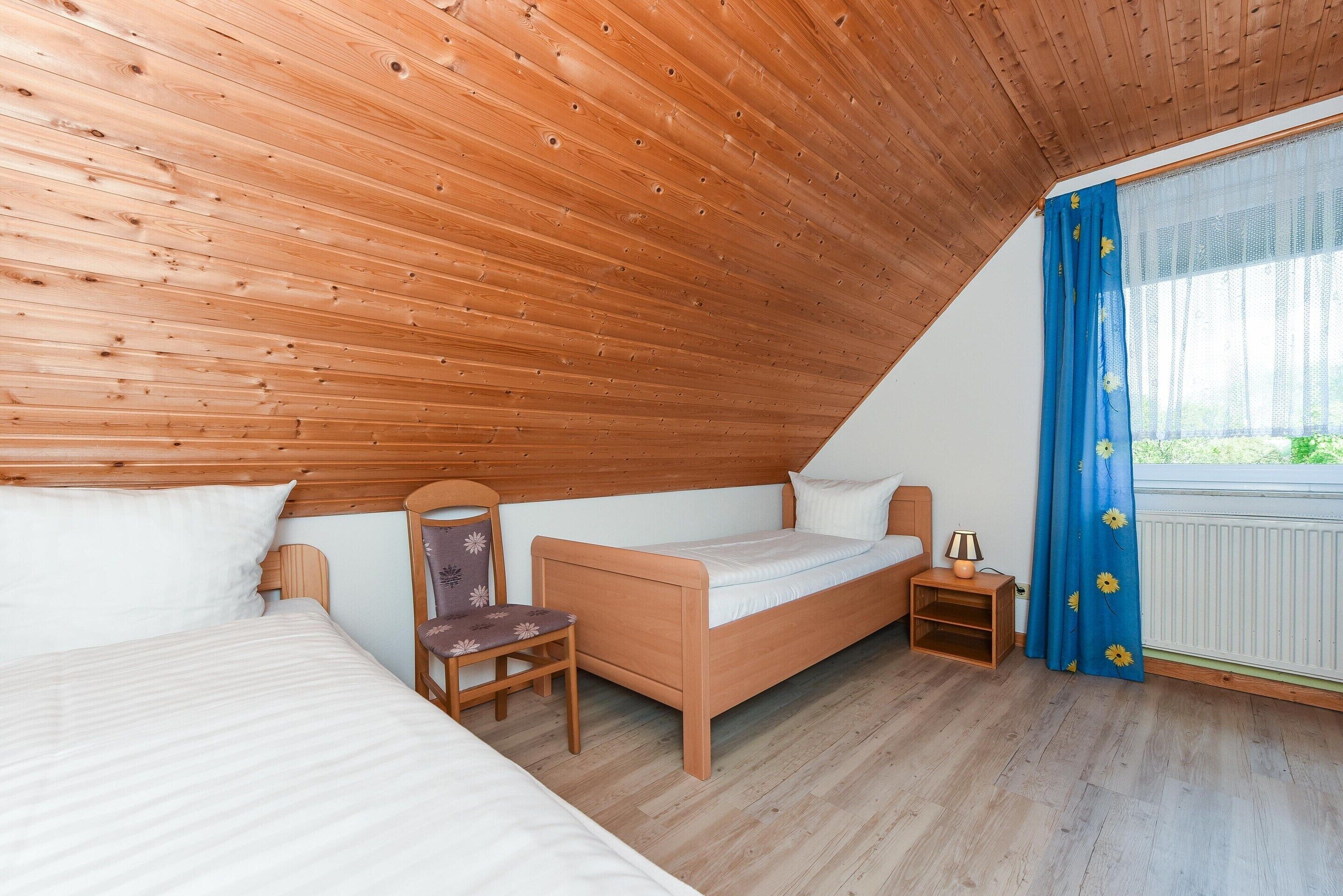 A cozy attic bedroom with a sloped ceiling, a wooden bed, a chair, a nightstand, and a window with blue curtains.
