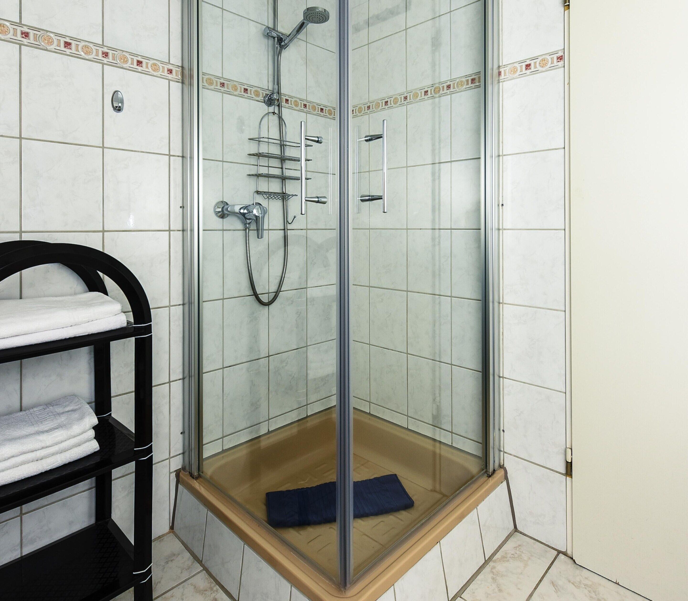 A glass-enclosed shower with a wooden base, tiled walls, and a black towel rack with folded towels.