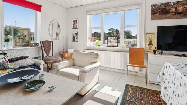 A well-lit living room with a sofa, armchairs, a coffee table, and a TV. Large windows provide natural light and a view of the outside.