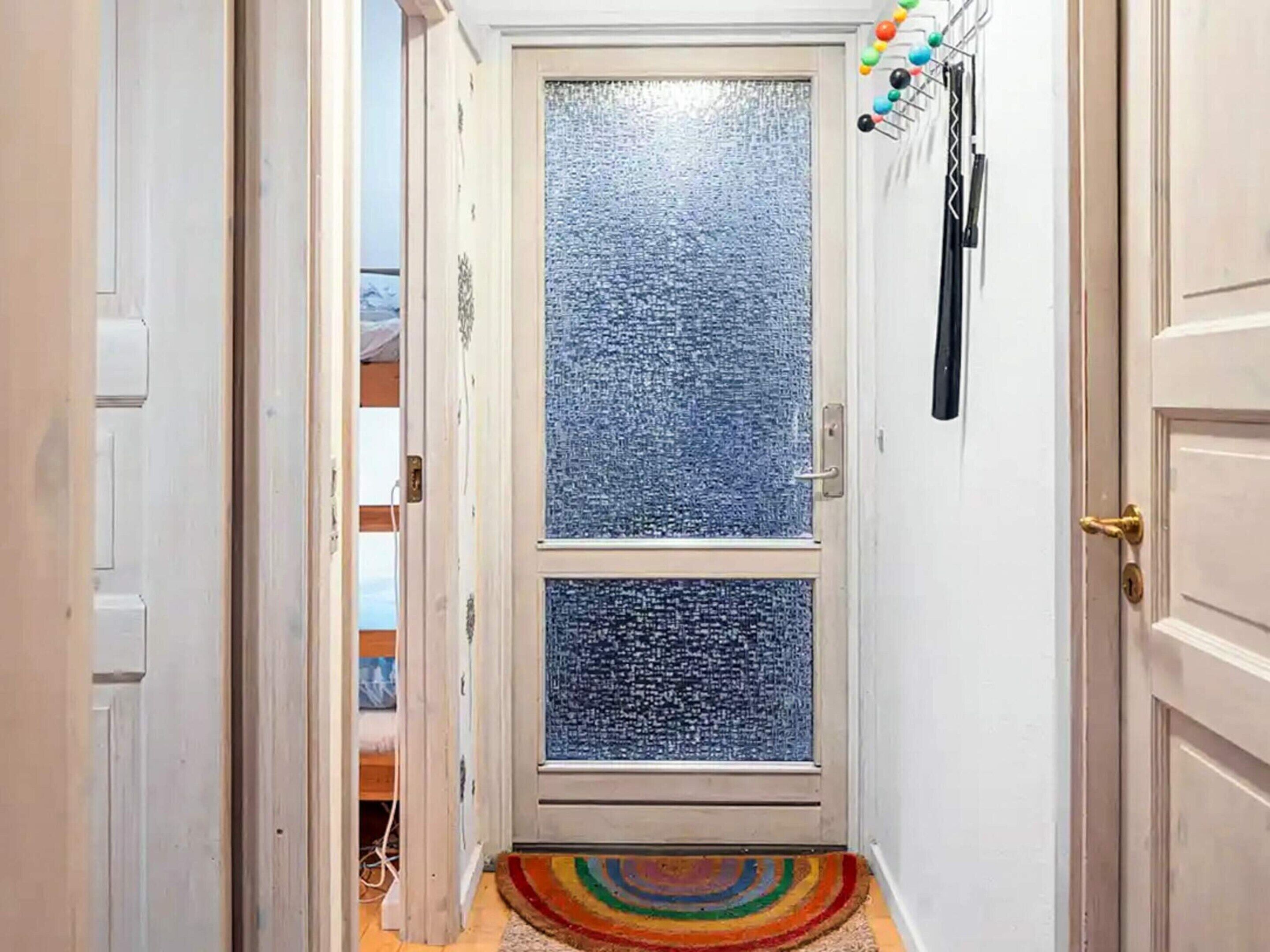 Een smalle gang met een deur van geëtst glas, een kleurrijk vloerkleed en een kapstok met jassen en accessoires.