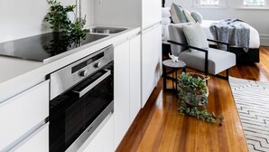 A modern kitchen with white cabinets, a stainless steel oven, and a sink. There is a small round table with a plant and a grey sofa in the adjacent living area. - Designer Styled Studio in Central Trendy Glebe (Sydney)