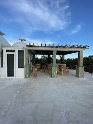 Et overdækket udendørs siddeområde med træbænk og pergola.