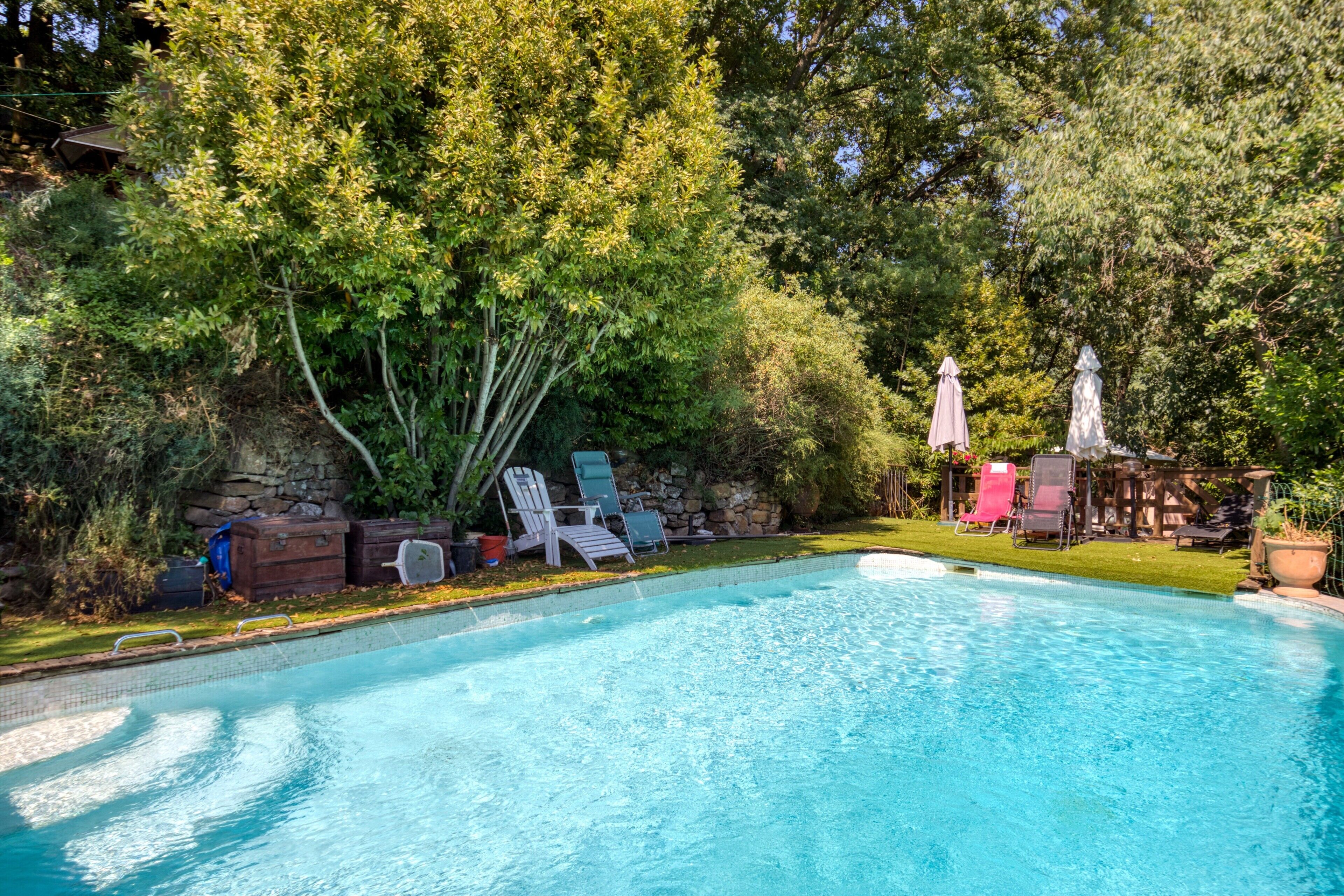 Una piscina con sillones de descanso y sombrillas, rodeada de árboles y un muro de piedra.