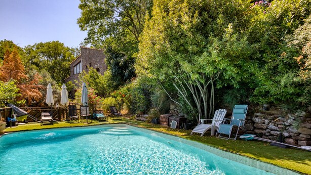 A swimming pool with clear water, surrounded by lounge chairs and umbrellas, with a stone wall and trees in the background. - Gite du bas – in La Rouquette, a charming little hamlet (Saint-Privat)