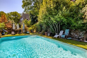 A swimming pool with clear water, surrounded by lounge chairs and umbrellas, with a stone wall and trees in the background. - Gite du bas – in La Rouquette, a charming little hamlet (Saint-Privat)