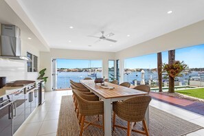 A modern kitchen with a view of the marina, a dining area with wicker chairs, and a ceiling fan. - Amazing Canal House With Private Jetty, Gym & Pool (Sunshine Coast)