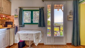A kitchen with wooden cabinets, a table with a floral tablecloth, a window with curtains, and a view of the outdoors. - Gite du haut – in La Rouquette, a charming little hamlet (Saint-Privat)