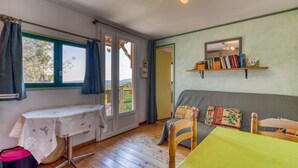 A room with a wooden floor, a table with a tablecloth, a sofa with cushions, a bookshelf, and a window with curtains. - Gite du haut – in La Rouquette, a charming little hamlet (Saint-Privat)