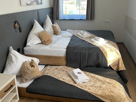 Ein Doppelbett mit grauer gesteppter Bettdecke und weißen Kissen, ein Nachttisch mit Schublade sowie ein Fenster mit Vorhängen.